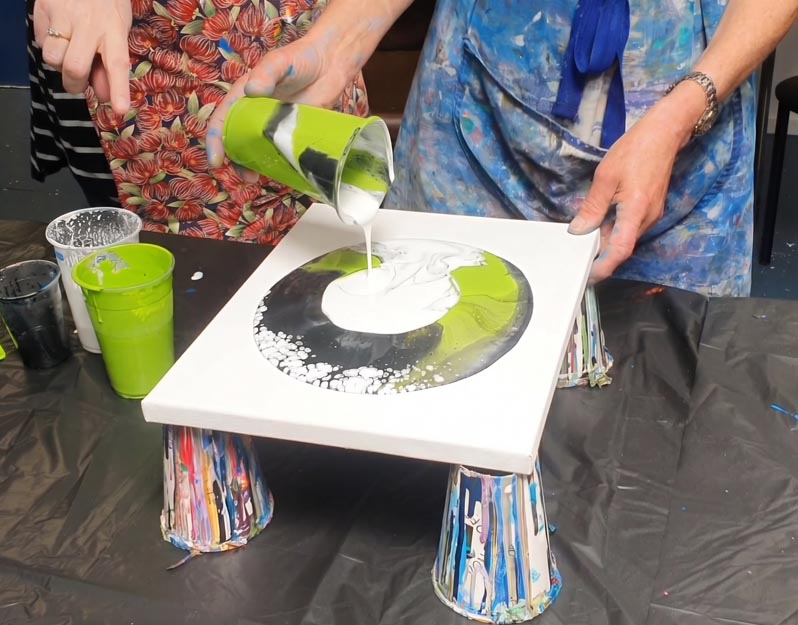 Hsnds pour white paint on to a canvas already with green and black paint on it